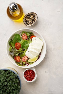 Yeşil yapraklı salata, marul, ıspanak, kiraz domatesleri, mozarella, zeytinyağı, kırmızı biber, gri taş yüzeyde limon suyu, üst manzara.