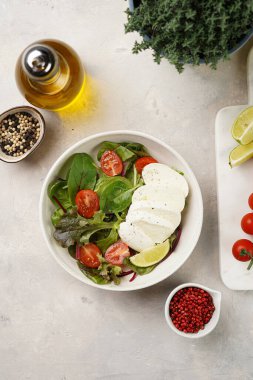 Yeşil yapraklı salata, marul, ıspanak, kiraz domatesleri, mozarella, zeytinyağı, kırmızı biber, gri taş yüzeyde limon suyu, üst manzara.