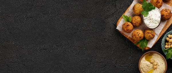 Geleneksel oryantal nohut, ahşap bir tahtada kızartılmış falafel, tzatziki yoğurt sosu, humus, siyah yüzey üzerinde taze kireç ve yeşil kişniş, kopyalama alanı, pankart.