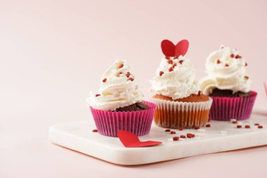 Beyaz kremalı üç ev yapımı beyaz ve çikolatalı kek ve pembe yüzeydeki mermer tahtada kalp paylaşımı kırmızı ve beyaz şeker süslemesi.