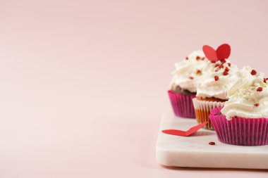 Beyaz kremalı üç ev yapımı beyaz ve çikolatalı kek ve pembe yüzeydeki mermer tahtada kalp paylaşımı kırmızı ve beyaz şeker süslemesi.