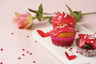 Kırmızı kremalı üç ev yapımı beyaz ve çikolatalı kek ve pembe yüzeydeki mermer tahtada kalp paylaşımı kırmızı ve beyaz şeker süslemesi.