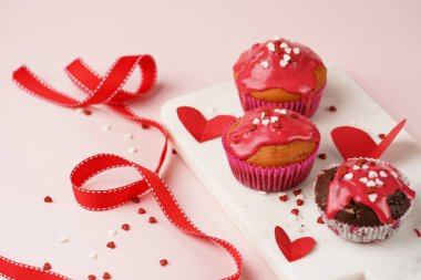 Kırmızı kremalı üç ev yapımı beyaz ve çikolatalı kek ve pembe yüzeydeki mermer tahtada kalp paylaşımı kırmızı ve beyaz şeker süslemesi.