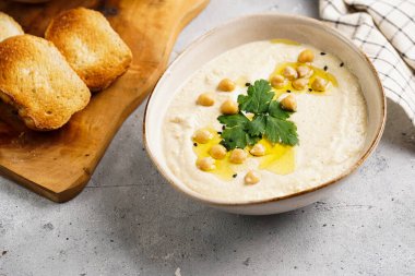 Nohut soslu humus ve maydanozlu seramik kase beton yüzeyde kızarmış baget dilimleri ve ahşap tahtada zeytinyağı.