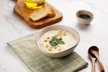 Nohut soslu humus ve maydanozlu seramik kasede mermer yüzeyde kızarmış baget dilimleri ve ahşap tahtada zeytinyağı.