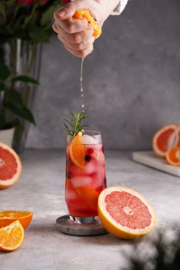 Kadın eli portakaldan meyve suyunu soğuk portakal suyu, greyfurt, donmuş böğürtlen ve buz küpleri, biberiye, greyfurt suyunu gri arka planda uzun bir bardağa sıkıyor.