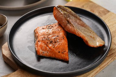 İki ızgara balık bifteği. Tahta tahtada siyah yuvarlak tabakta kırmızı somon, zeytinyağı, gri arka planda avokado.