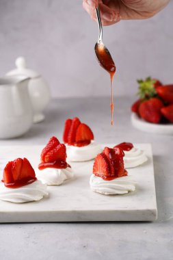 Taze çilekli mini pavlovanın üzerine kırmızı reçel dökmek. Mermer tahtada, beyaz arka planda lezzetli kremalı pastalar.