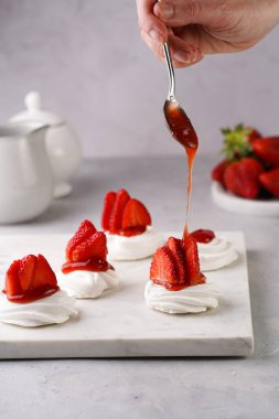 Taze çilekli mini pavlovanın üzerine kırmızı reçel dökmek. Mermer tahtada, beyaz arka planda lezzetli kremalı pastalar.
