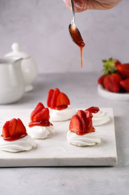 Taze çilekli mini pavlovanın üzerine kırmızı reçel dökmek. Mermer tahtada, beyaz arka planda lezzetli kremalı pastalar.