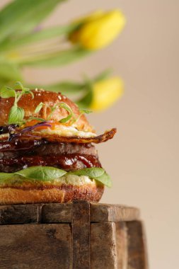 Duble angus bifteği köfteli hamburger, domates, ıspanak, sahanda yumurta ve barbekü sosu ahşap tahtada, pastel arka planda gerçek lalelerle.