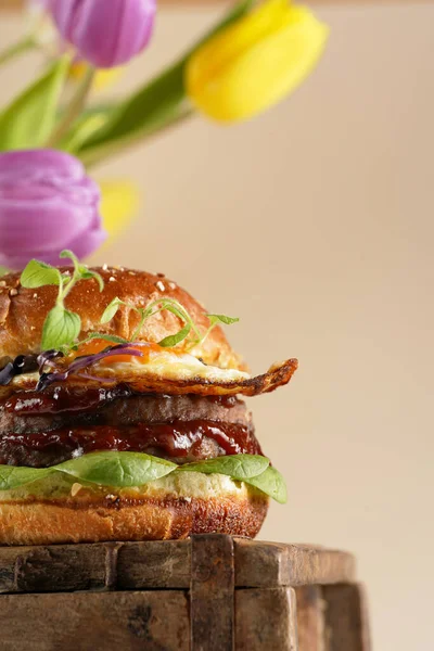 Duble angus bifteği köfteli hamburger, domates, ıspanak, sahanda yumurta ve barbekü sosu ahşap tahtada, pastel arka planda gerçek lalelerle.