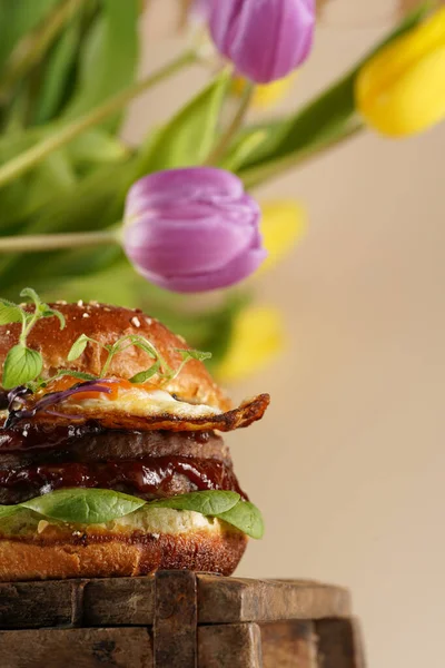 Duble angus bifteği köfteli hamburger, domates, ıspanak, sahanda yumurta ve barbekü sosu ahşap tahtada, pastel arka planda gerçek lalelerle.