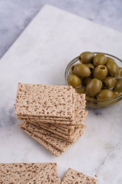 Sağlıklı çıtır çıtır krakerler, susam tohumları ve mermer tahtada yeşil zeytinler.
