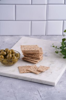 Sağlıklı çıtır çıtır krakerler, susam tohumları ve mermer tahtada yeşil zeytinler.
