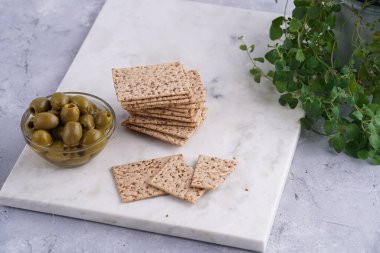Sağlıklı çıtır çıtır krakerler, susam tohumları ve mermer tahtada yeşil zeytinler.