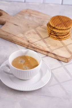 Stroopwafel - geleneksel Hollanda usulü karamelli gözleme - tahtanın üzerinde üst üste istiflenmiş beyaz mutfak ve beyaz porselen fincanda bir fincan kahve