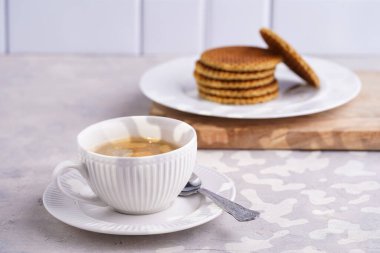 Stroopwafel - geleneksel Hollanda usulü karamelli gözleme - tahtanın üzerinde üst üste istiflenmiş beyaz mutfak ve beyaz porselen fincanda bir fincan kahve