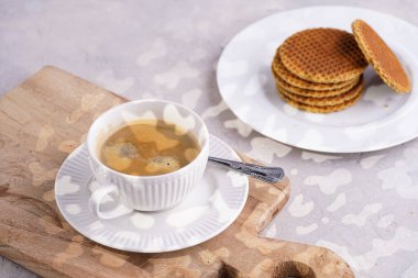 Stroopwafel - geleneksel Hollanda usulü karamelli gözleme - tahtanın üzerinde üst üste istiflenmiş beyaz mutfak ve beyaz porselen fincanda bir fincan kahve