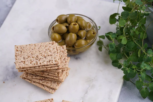 Sağlıklı çıtır çıtır krakerler, susam tohumları ve mermer tahtada yeşil zeytinler.