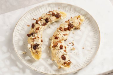 Fıstık ezmesi soslu iki muz, kuru üzüm, badem ve beyaz tabakta chia tohumları mermer tahtada, doğrudan yaratıcı güneş ışığı gölgesi