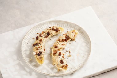 Fıstık ezmesi soslu iki muz, kuru üzüm, badem ve beyaz tabakta chia tohumları mermer tahtada, doğrudan yaratıcı güneş ışığı gölgesi