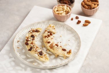 Fıstık ezmesi soslu iki muz, kuru üzüm, badem ve beyaz tabakta chia tohumları mermer tahtada, doğrudan yaratıcı güneş ışığı gölgesi