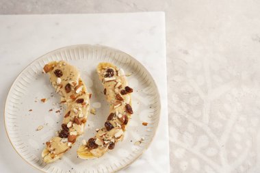 Fıstık ezmesi soslu iki muz, kuru üzüm, badem ve beyaz tabakta Chia tohumları mermer tahtada, doğrudan yaratıcı güneş ışığı gölgesi. en üstteki görünüm
