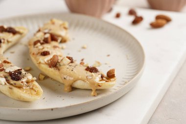 Fıstık ezmesi soslu iki muz, kuru üzüm, badem ve beyaz tabakta Chia tohumları mermer tahtada, doğrudan yaratıcı güneş ışığı gölgesi. Fıstık ezmesine yakın çekim