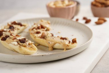 Fıstık ezmesi soslu iki muz, kuru üzüm, badem ve beyaz tabakta chia tohumları mermer tahtada, doğrudan yaratıcı güneş ışığı gölgesi