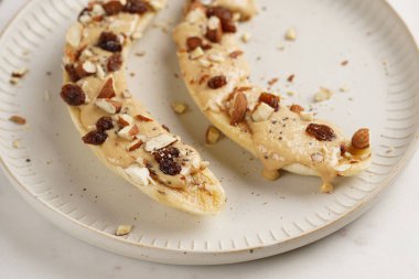 Fıstık ezmesi soslu iki muz, kuru üzüm, badem ve beyaz tabakta chia tohumları mermer tahtada, doğrudan yaratıcı güneş ışığı gölgesi