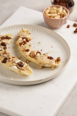 Fıstık ezmesi soslu iki muz, kuru üzüm, badem ve beyaz tabakta chia tohumları mermer tahtada, doğrudan yaratıcı güneş ışığı gölgesi