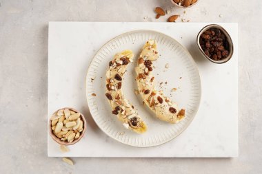 Fıstık ezmesi soslu iki muz, kuru üzüm, badem ve beyaz tabakta Chia tohumları mermer tahtada, doğrudan yaratıcı güneş ışığı gölgesi. en üstteki görünüm