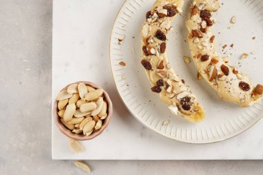 Fıstık ezmesi soslu iki muz, kuru üzüm, badem ve beyaz tabakta Chia tohumları mermer tahtada, doğrudan yaratıcı güneş ışığı gölgesi. en üstteki görünüm