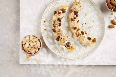 Fıstık ezmesi soslu iki muz, kuru üzüm, badem ve beyaz tabakta Chia tohumları mermer tahtada, doğrudan yaratıcı güneş ışığı gölgesi. en üstteki görünüm