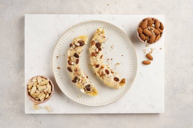 Fıstık ezmesi soslu iki muz, kuru üzüm, badem ve beyaz tabakta Chia tohumları mermer tahtada, doğrudan yaratıcı güneş ışığı gölgesi. en üstteki görünüm