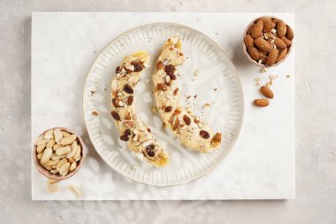 Fıstık ezmesi soslu iki muz, kuru üzüm, badem ve beyaz tabakta Chia tohumları mermer tahtada, doğrudan yaratıcı güneş ışığı gölgesi. en üstteki görünüm