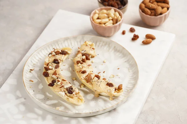 Fıstık ezmesi soslu iki muz, kuru üzüm, badem ve beyaz tabakta chia tohumları mermer tahtada, doğrudan yaratıcı güneş ışığı gölgesi