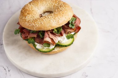 Pastırmalı simit sandviç, salatalık dilimleri, su teresi salatası ve mermer zemin üzerinde ricotta.