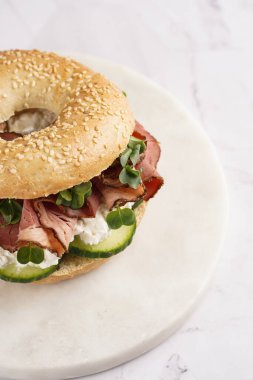 Pastırmalı simit sandviç, salatalık dilimleri, su teresi salatası ve mermer zemin üzerinde ricotta.