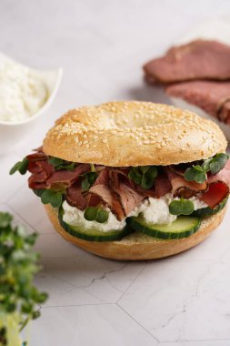 Pastırmalı simit sandviç, salatalık dilimleri, su teresi salatası ve mermer zemin üzerinde ricotta.