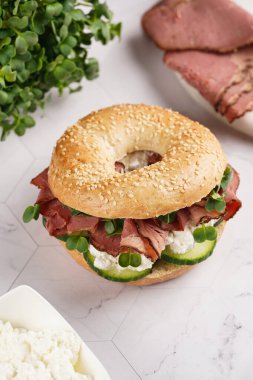 Pastırmalı simit sandviç, salatalık dilimleri, su teresi salatası ve mermer arka planda ricotta.