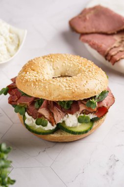 Pastırmalı simit sandviç, salatalık dilimleri, su teresi salatası ve mermer zemin üzerinde ricotta.