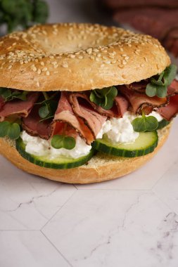 Pastırmalı simit sandviç, salatalık dilimleri, su teresi salatası ve mermer zemin üzerinde ricotta.