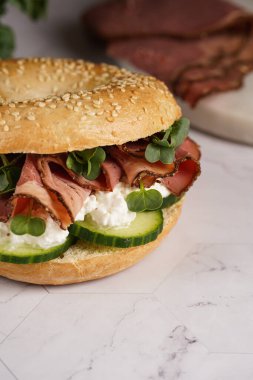 Pastırmalı simit sandviç, salatalık dilimleri, su teresi salatası ve mermer zemin üzerinde ricotta.