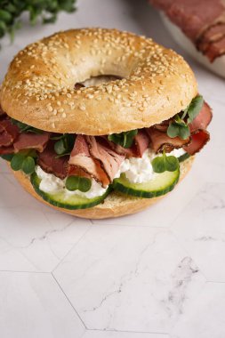 Pastırmalı simit sandviç, salatalık dilimleri, su teresi salatası ve mermer zemin üzerinde ricotta.