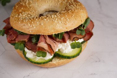 Pastırmalı simit sandviç, salatalık dilimleri, su teresi salatası ve mermer zemin üzerinde ricotta.