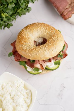 Pastırmalı simit sandviç, salatalık dilimleri, su teresi salatası ve mermer zemin üzerinde ricotta.