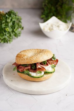 Pastırmalı simit sandviç, salatalık dilimleri, su teresi salatası ve mermer zemin üzerinde ricotta.