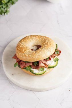 Pastırmalı simit sandviç, salatalık dilimleri, su teresi salatası ve mermer zemin üzerinde ricotta.